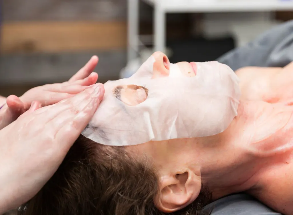Hands applying a sheet mask during a facial at Piedmont Injury Massage in Hoschton, GA