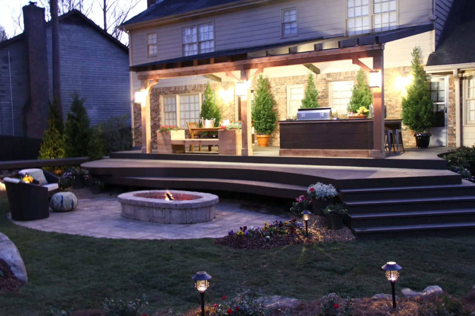 Illuminated pergola, furnished deck, and circular fire pit at dusk