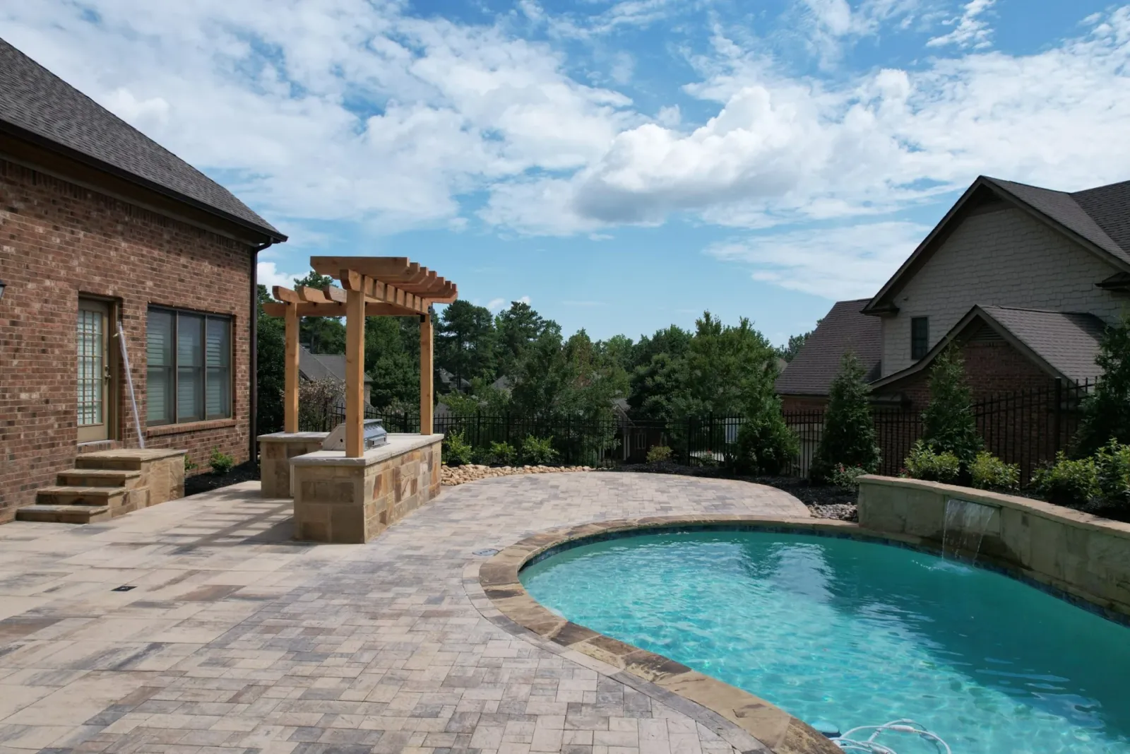 Poolside outdoor kitchen, stone patio, and timber pergola beside a brick home
