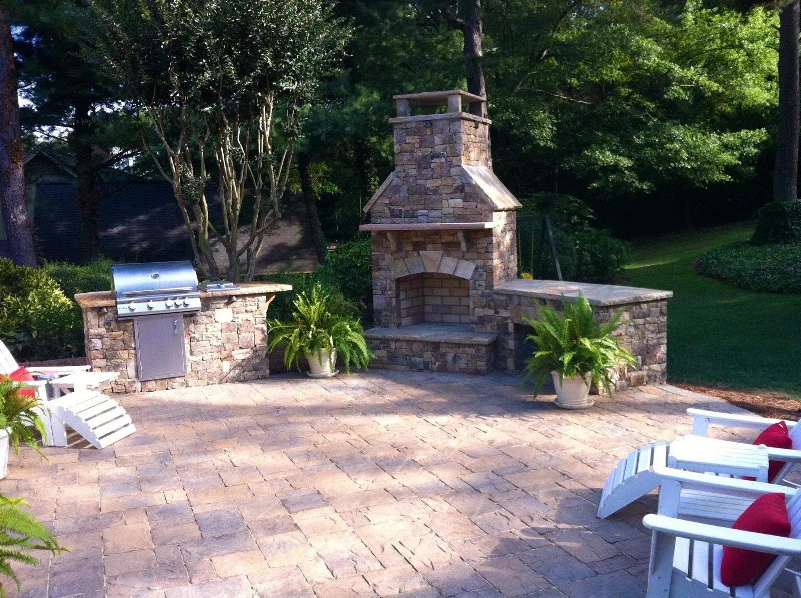 Stone outdoor fireplace and grill island with patio seating beside a wooded lawn