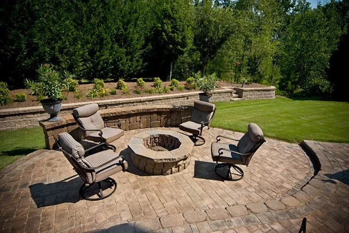 Circular paver patio with a stone fire pit, lounge chairs, and a seating wall