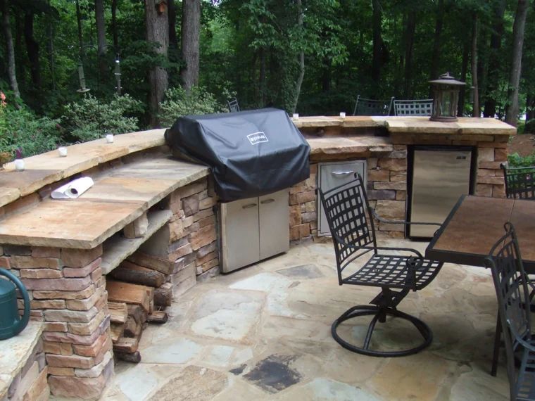 Curved stone outdoor kitchen with a grill, countertop seating, and a wooded backdrop