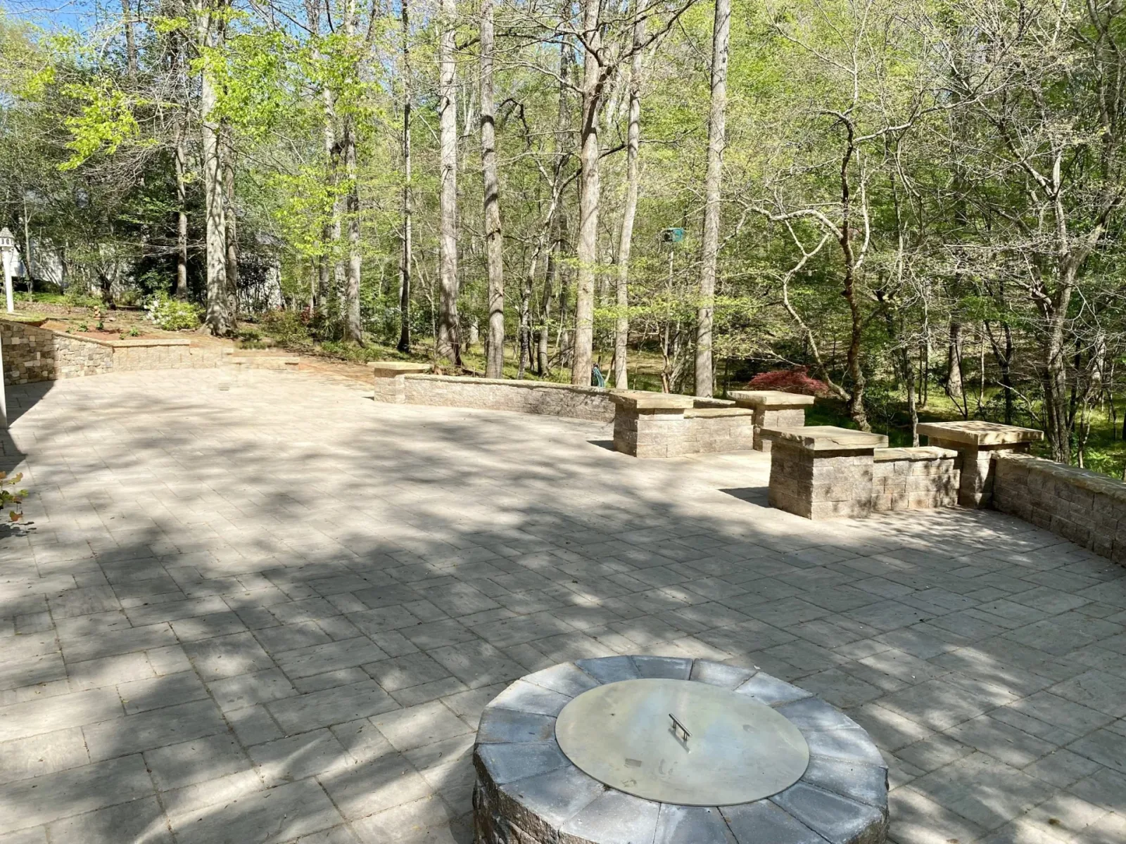 Broad paver patio, curved stone walls, and a fire pit beside a wooded garden