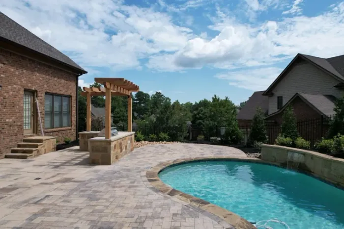 Swimming pool and broad stone pool deck beside an outdoor kitchen and pergola